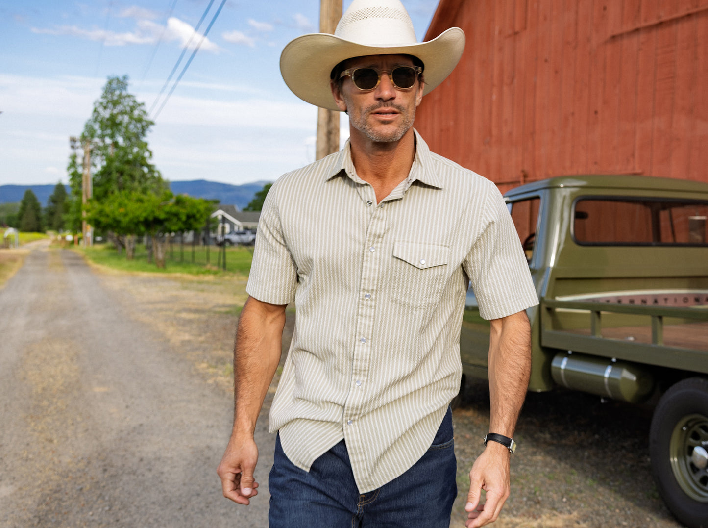 Man in a cowboy hat and sunglasses walking on a dirt road with a red barn and green truck in the background.
