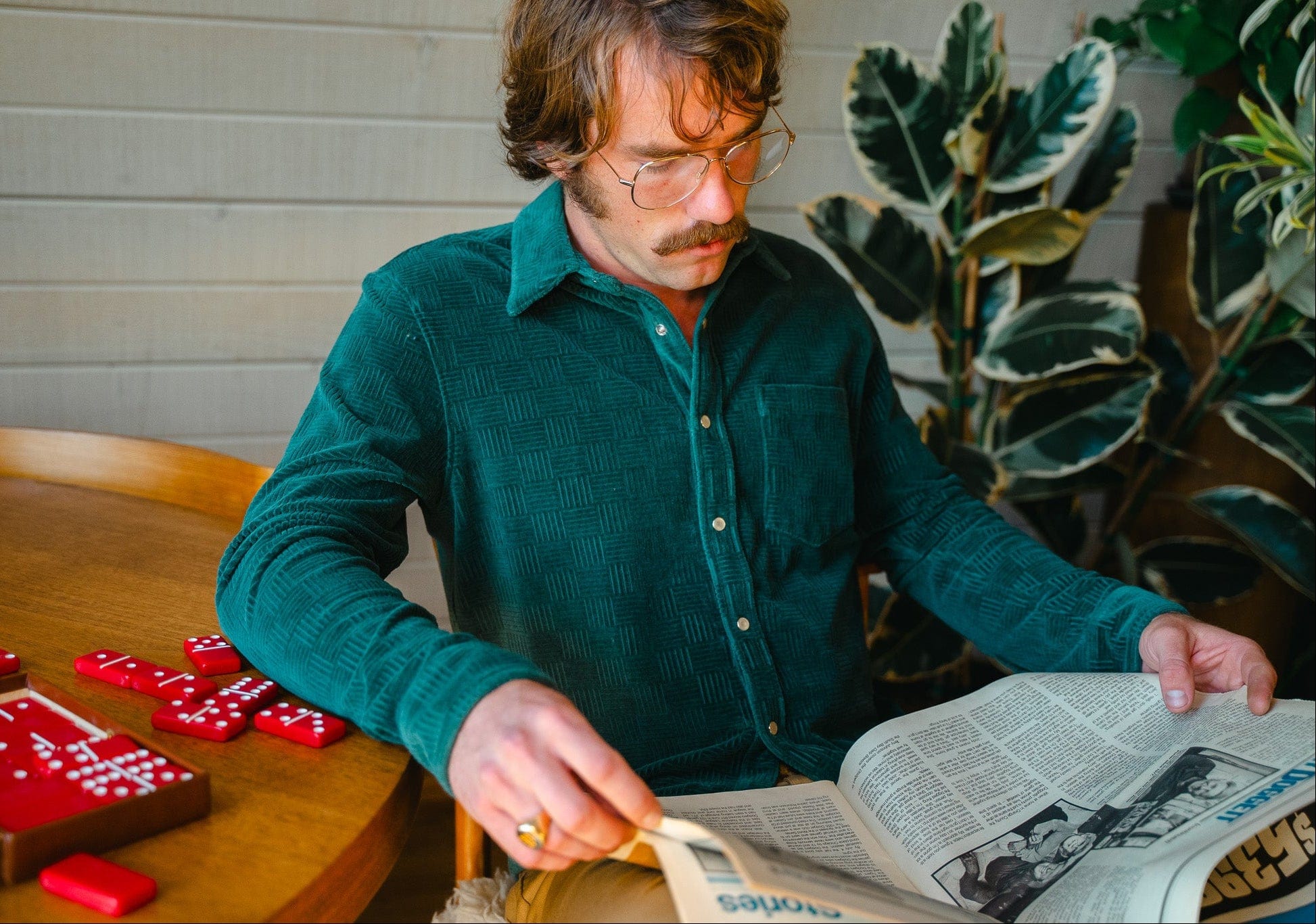 Man in a green shirt reading a newspaper at a table with dice