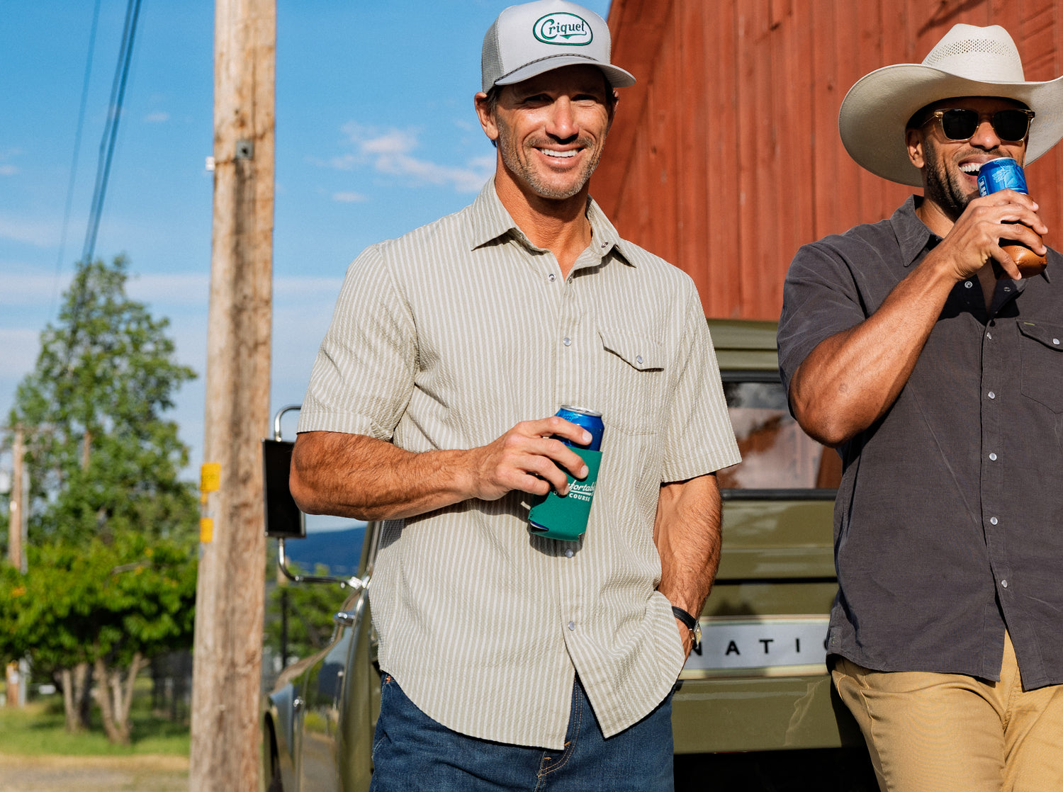 Two men standing outdoors, one holding a blue cup and the other a can, with a red barn in the background.
