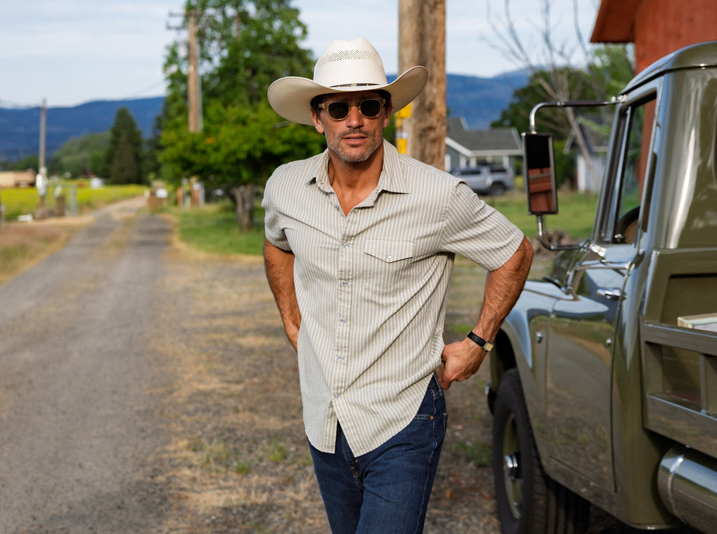 Man in cowboy hat and sunglasses standing next to a truck on a rural road.