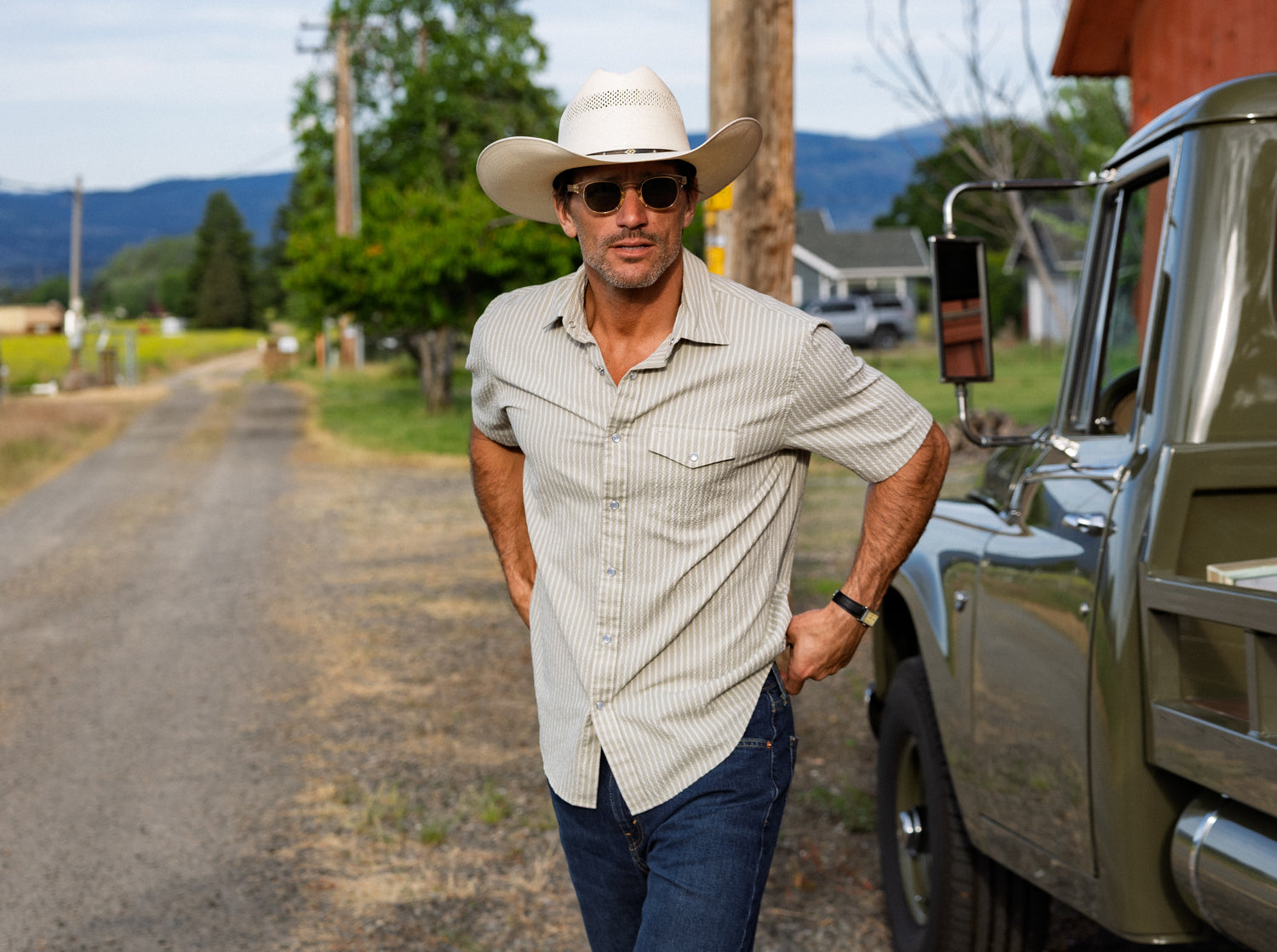Man in cowboy hat and sunglasses standing next to a truck on a rural road.