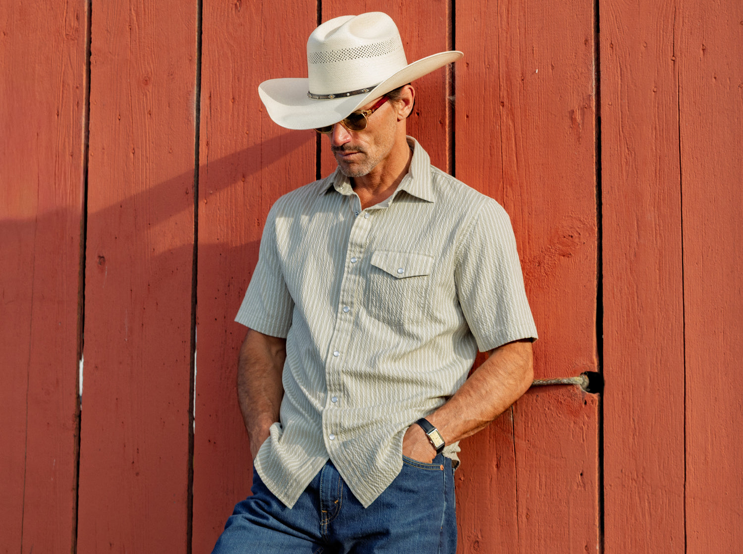 Man wearing a cowboy hat and sunglasses leaning against a red wooden wall.