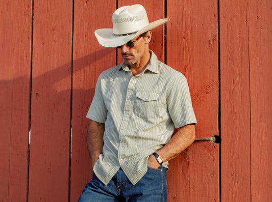 Man wearing a cowboy hat and sunglasses leaning against a red wooden wall.